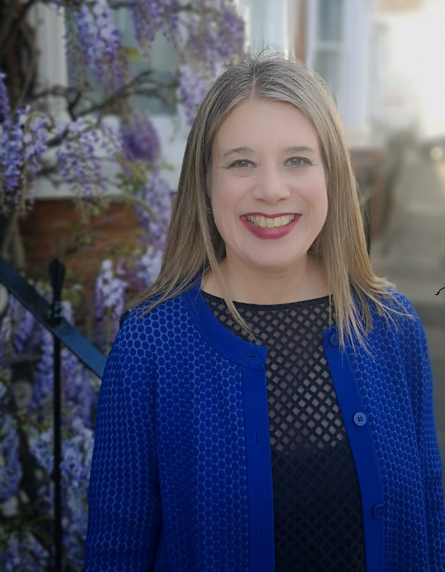 Dawn_Reflexologist Photo of Dawn, Reflexologist, wearing a bright blue jacket, stood in front of purple wisteria